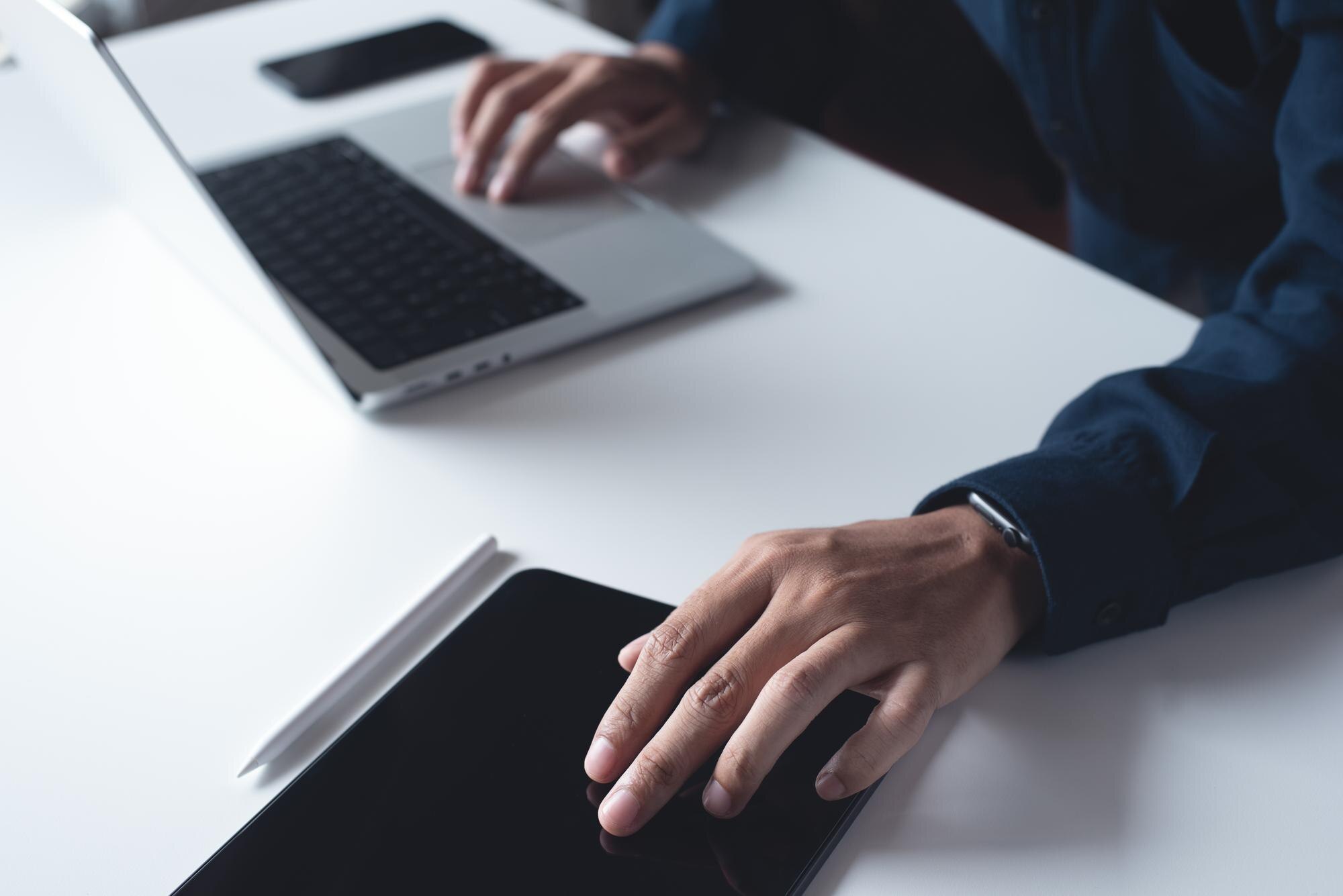 Mørk dresshånd bruker en digital tablett mens den andre er på en laptop. En mobiltelefon ligger på bordet.