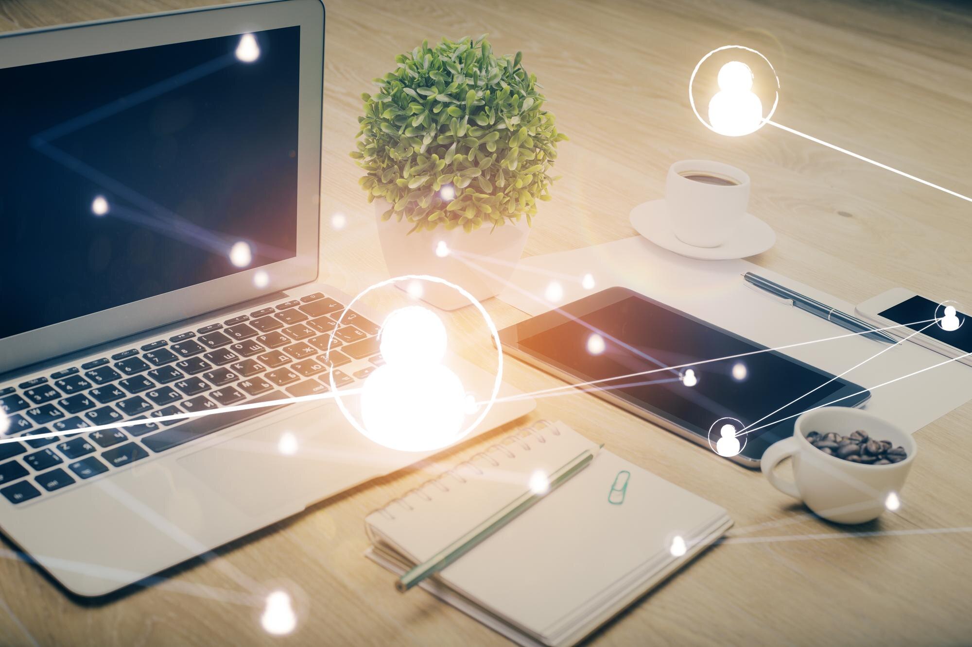 Laptopen er åpen på et skrivebord med en plante, en kopp kaffe, en notisbok og et nettbrett, omgitt av hologram-lignende sosiale nettverksikoner.