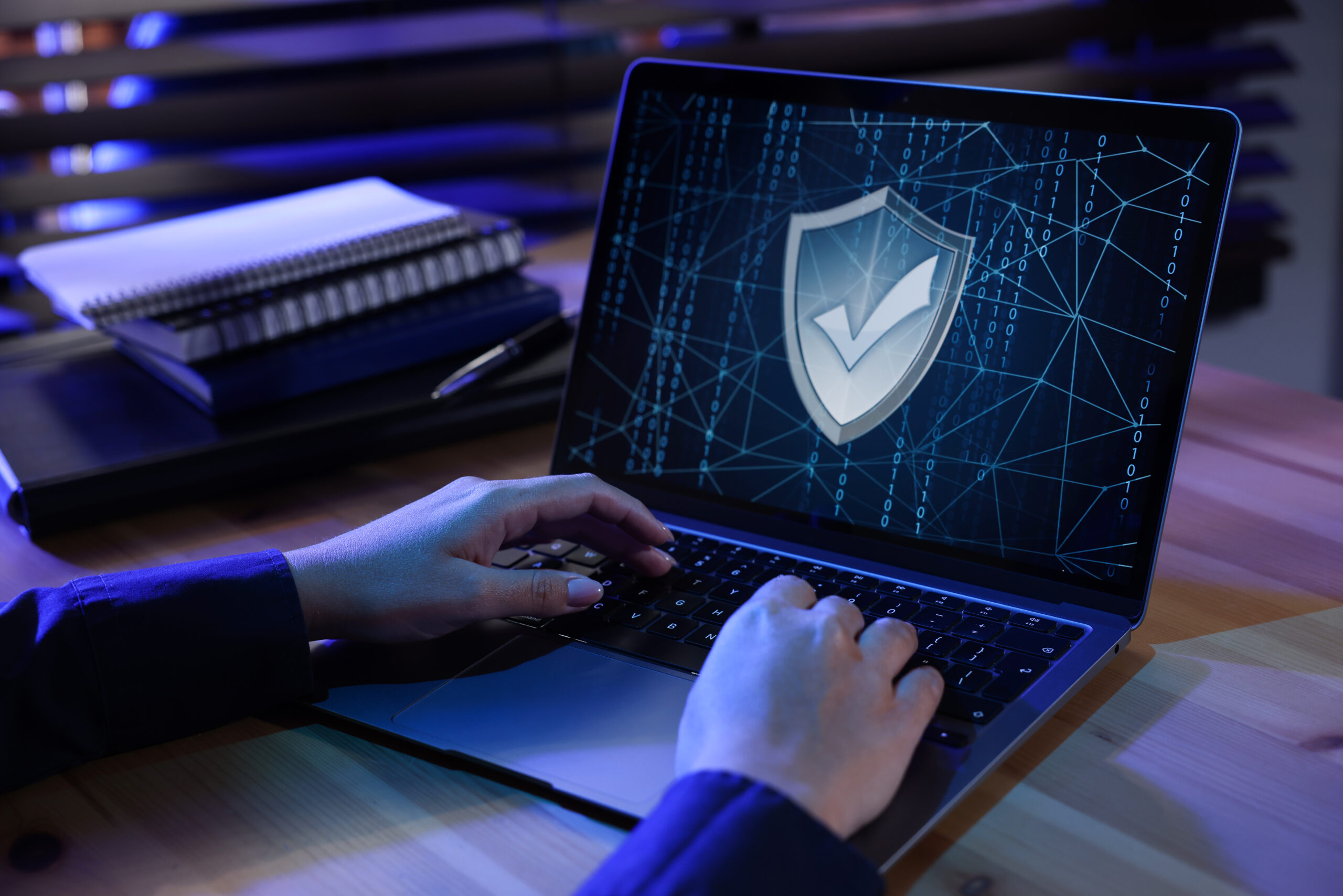 En person som skriver på en bærbar datamaskin med et digitalt skjold med en hake, som symboliserer cybersikkerhet eller databeskyttelse, med en notatbok og penn på skrivebordet i et svakt opplyst rom. En person som skriver på en bærbar datamaskin med et digitalt skjold med en hake, som symboliserer cybersikkerhet eller databeskyttelse, med en notatbok og penn på skrivebordet i et svakt opplyst rom.