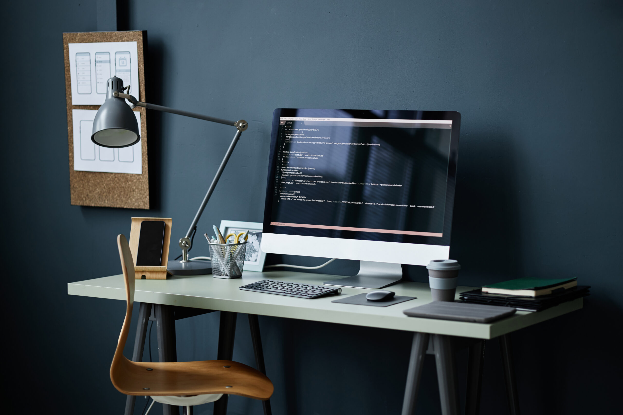 En moderne arbeidsplass med en datamaskin som viser kode, en skrivebordslampe, en kaffekopp, notatbøker og annet kontorrekvisita på et lyst skrivebord mot en mørk vegg, med skisser festet på en korktavle i bakgrunnen. Regelmessig nettsidevedlikehold sikrer stabil drift, optimal ytelse og sikkerhet, beskytter mot tap av kunder og data. Web Design AS tilbyr skreddersydde planer og gratis konsultasjon.