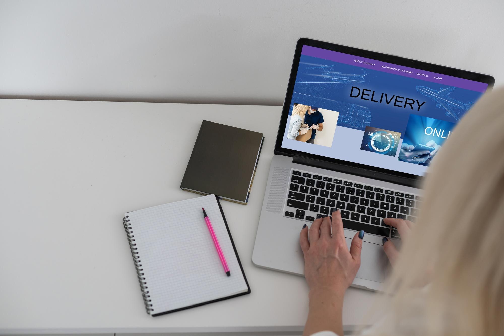 Kvinnelige hender på en bærbar datamaskin som viser en nettside med teksten "DELIVERY". Notatblokk og penn ligger ved siden av.
