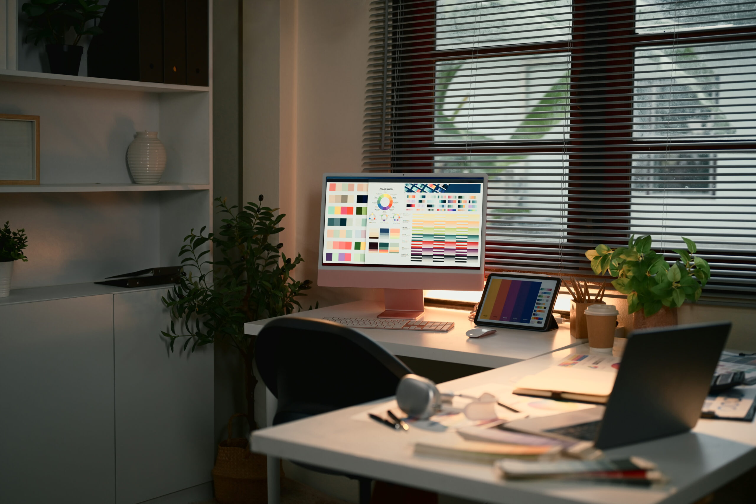 Titler: Moderne grafisk designarbeidsplass med planter Sammendrag: Stilig arbeidsmiljø for grafisk design med teknologi og planter. Lyst kontor med en stor skjerm, et laptop og planter, omgitt av naturlig lys som strømmer inn gjennom persienner.