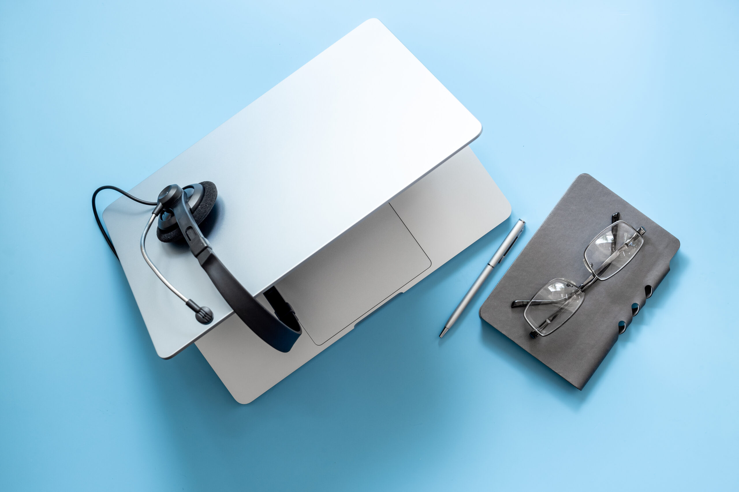 Kundestøtteutstyr med hodetelefoner og laptop Hodetelefoner, laptop og notatbok på blå bakgrunn. Sølv laptop med svart headset, en blyant og briller ved siden av på et blått bord.