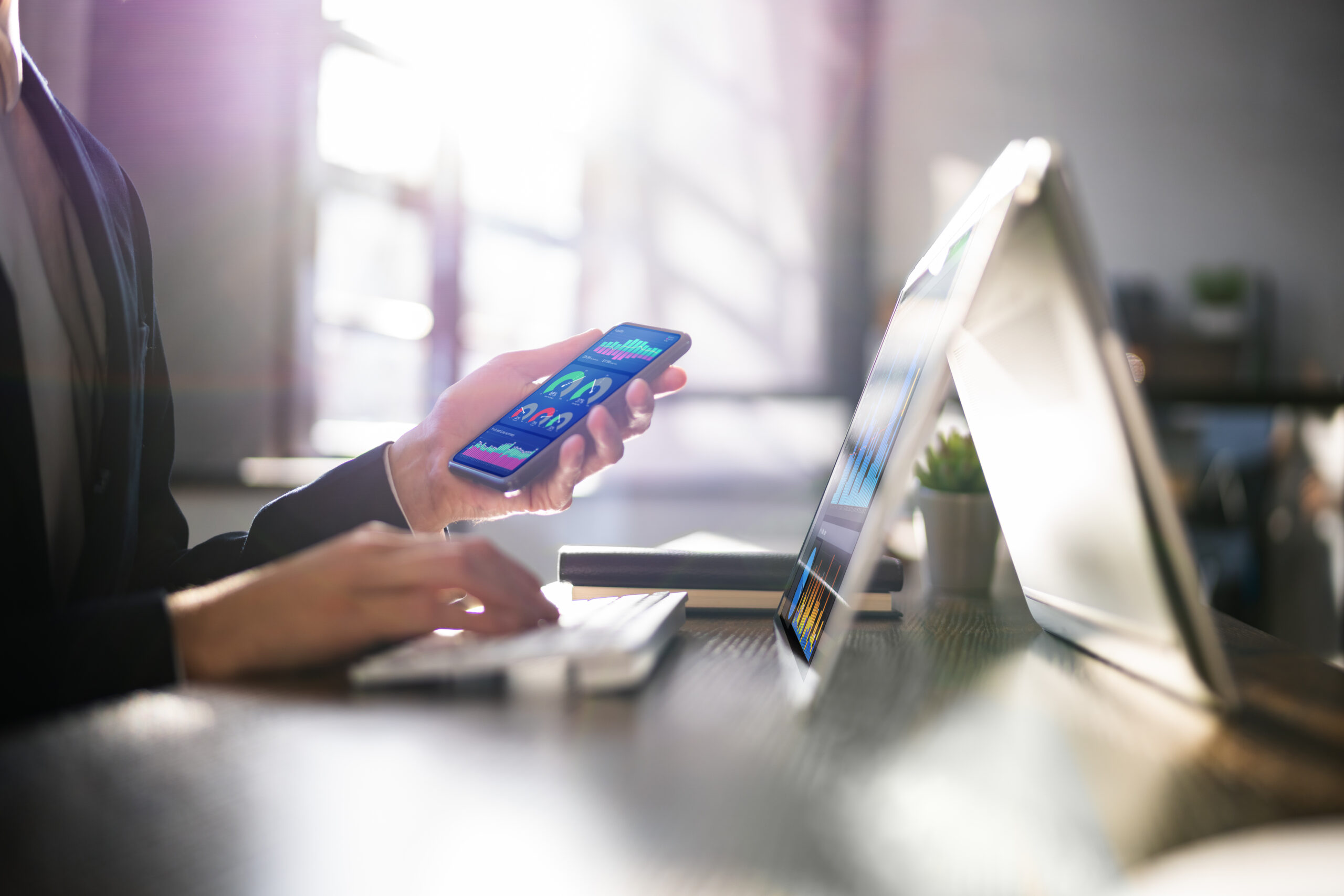 En person holder en smarttelefon i venstre hånd, mens høyre hånd skriver på et laptop-tastatur. Lys strømmer inn fra vinduet.