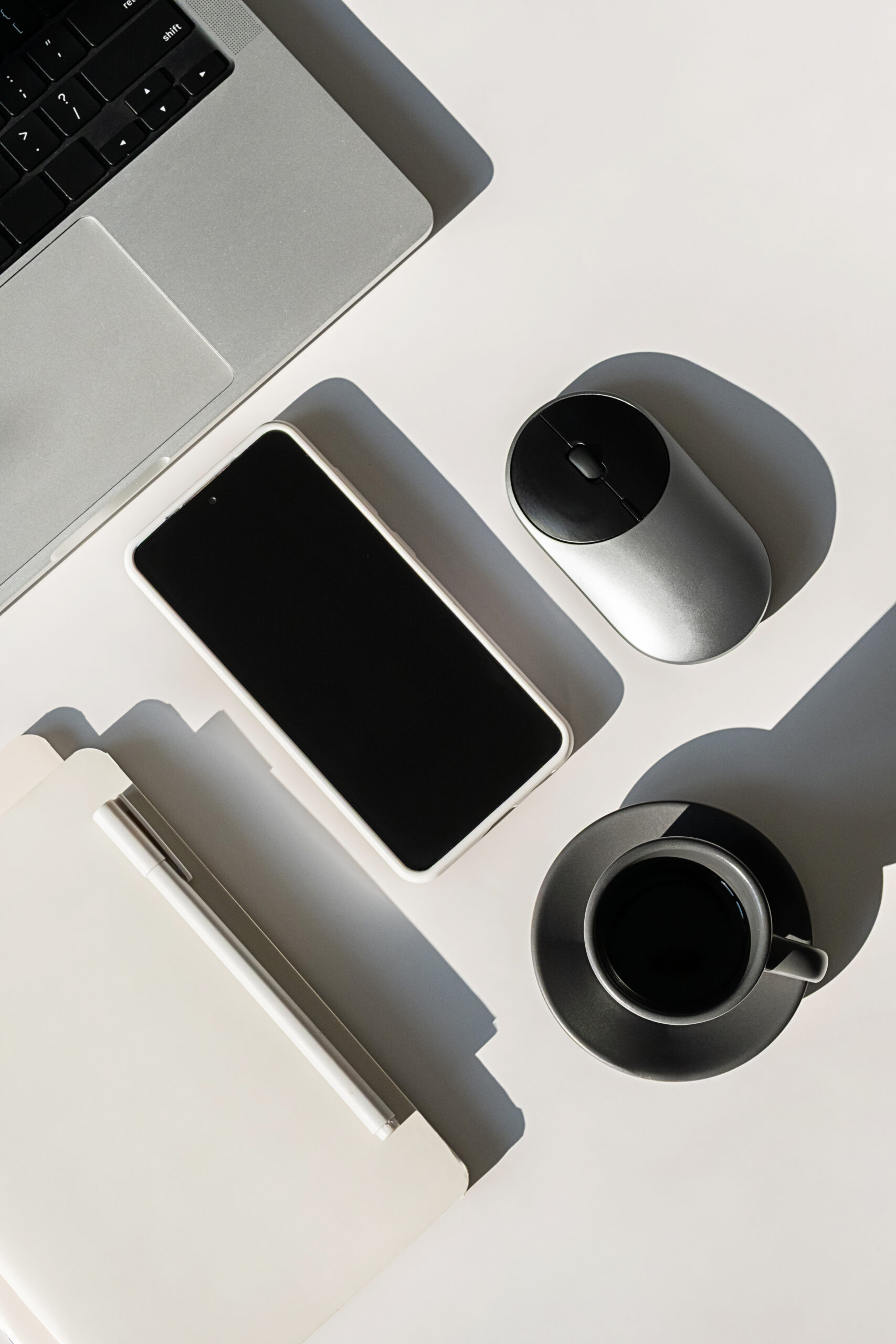 En bærbar datamaskin, en trådløs mus, en smarttelefon, et utklippstavle med papirer og en kopp svart kaffe er sirlig plassert på en hvit flate med skygger fra gjenstandene.