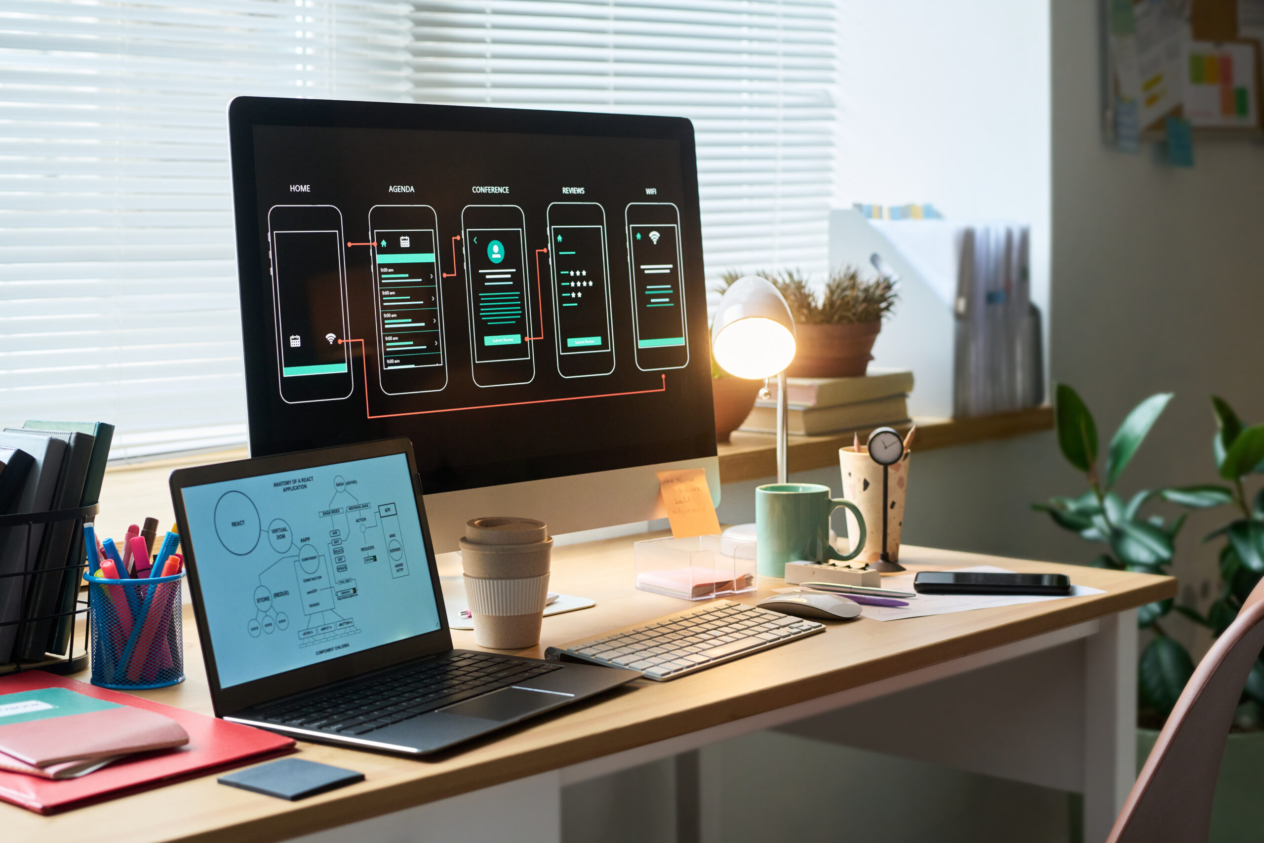 Moderne kontor med en stor skjerm som viser mobilappdesign, en laptop, skriveverktøy, planter og en lampe på arbeidsbordet.