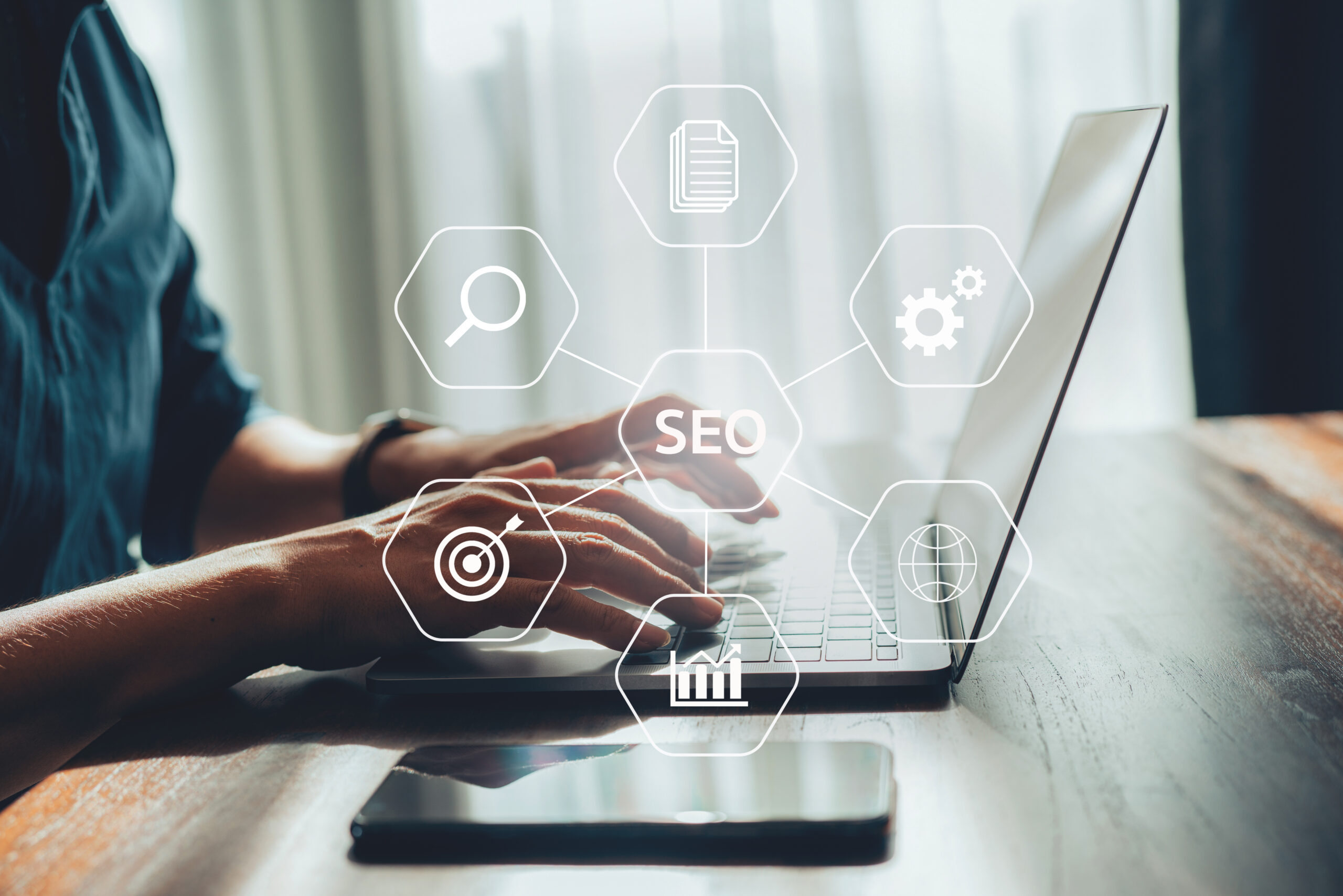 SEO-strategi: Optimalisering av søkemotorer i digital markedsføring Bilde av en person som jobber med SEO på datamaskin. Hender skriver på en laptop, omgitt av ikoner som representerer SEO-begreper, med en smarttelefon på bordet.