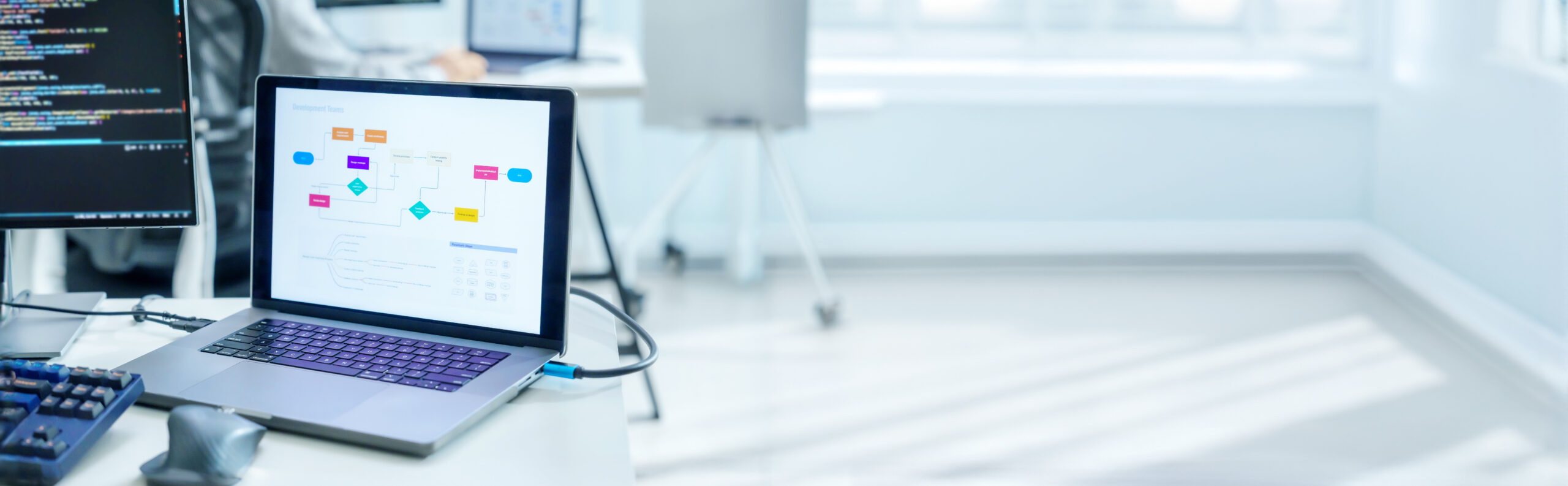 En bærbar datamaskin med et fargerikt flytskjema står på et skrivebord ved siden av en skjerm med kode i et lyst, moderne kontor. Et staffeli og store vinduer er synlige i bakgrunnen.