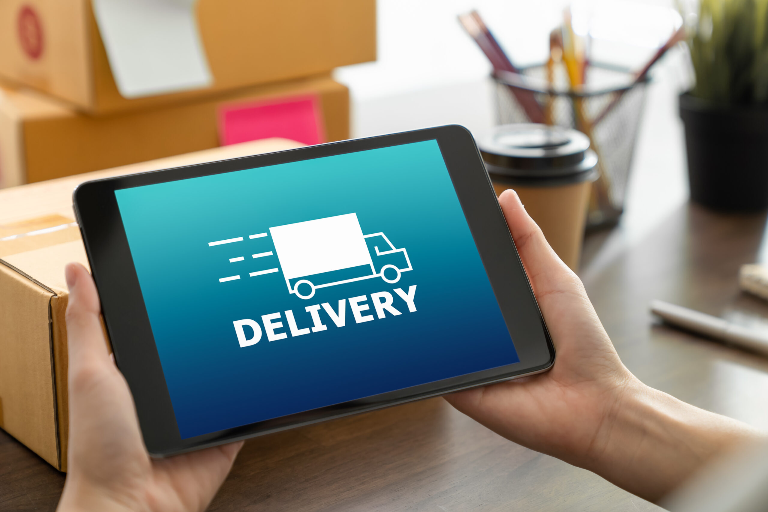 Hender som holder et nettbrett med "DELIVERY"-symbol, omgitt av pakker, en kaffe og kontorutstyr på et bord.