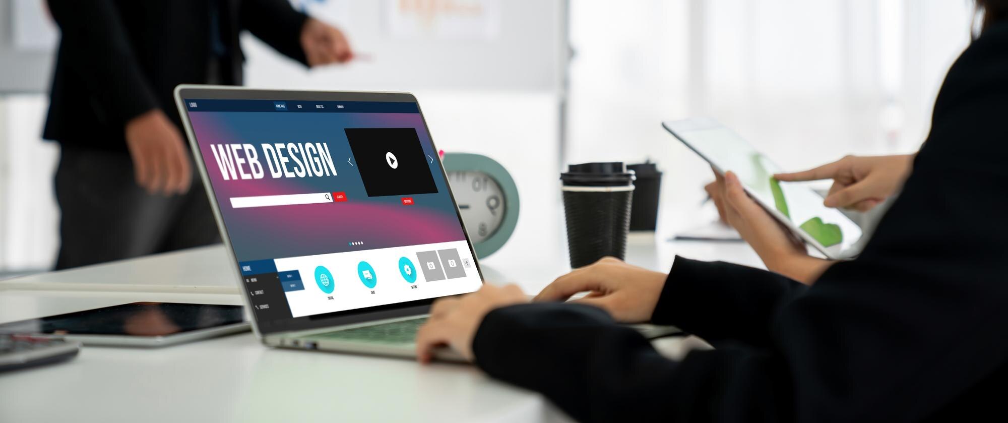 Laptopen viser et webdesign-grensesnitt med fargerike ikoner og teksten "WEB DESIGN". Kaffe og en mobiltelefon er synlige på bordet.